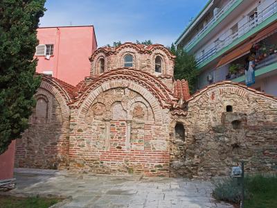 The image shows the exterior of the Byzantine Baths of Thessaloniki, a historic brick and stone structure with arched windows and terracotta roofing, situated between a pink building and a modern apartment block.