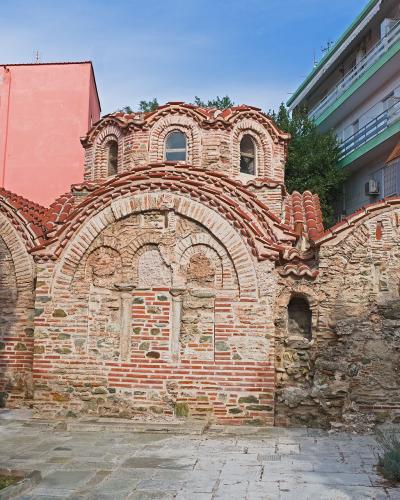 The image shows the exterior of the Byzantine Baths of Thessaloniki, a historic brick and stone structure with arched windows and terracotta roofing, situated between a pink building and a modern apartment block.