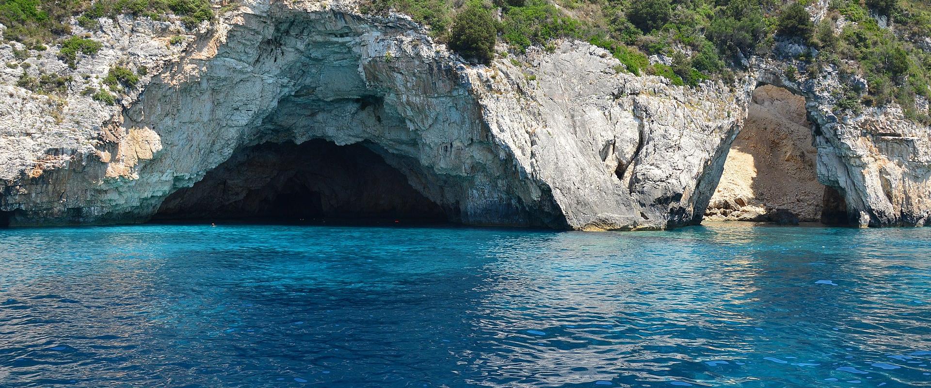A view of sea caves with bright blue water at the base of a limestone cliff covered in green vegetation.