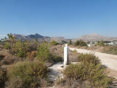 A white stone pillar with a spherical top stands in a dry, scrubby landscape with mountains in the background and a dirt road nearby.