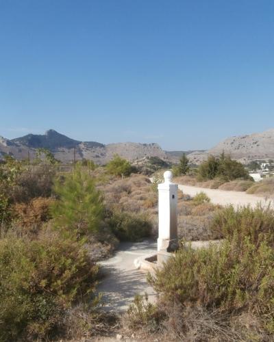 A white stone pillar with a spherical top stands in a dry, scrubby landscape with mountains in the background and a dirt road nearby.