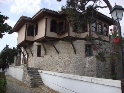 A two-story historic building with a stone lower level and a wooden upper story featuring overhanging balconies and dark shutters, situated next to a white wall and a lamppost.