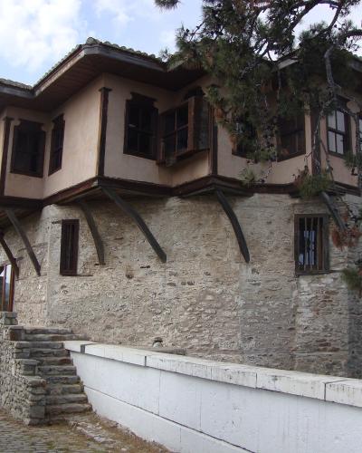 A two-story historic building with a stone lower level and a wooden upper story featuring overhanging balconies and dark shutters, situated next to a white wall and a lamppost.