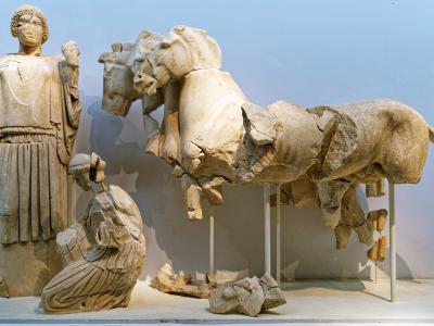 A museum interior displaying ancient Greek marble sculptures, including a standing female figure (Hera), a kneeling figure, and a large fragment of a quadriga (four-horse chariot) supported by metal stands.