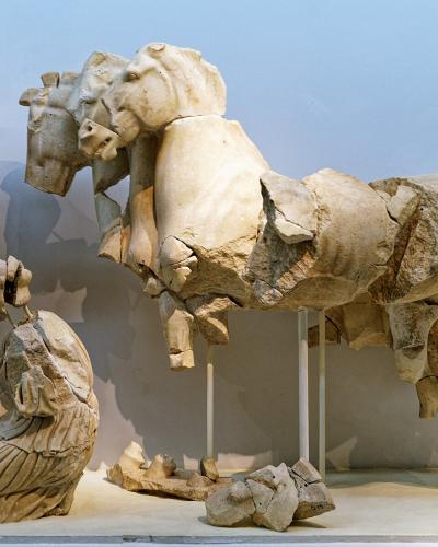 A museum interior displaying ancient Greek marble sculptures, including a standing female figure (Hera), a kneeling figure, and a large fragment of a quadriga (four-horse chariot) supported by metal stands.