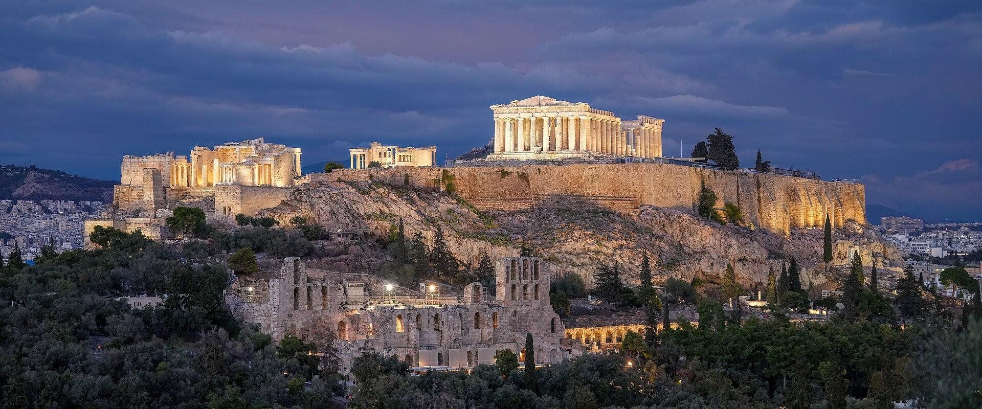 A night photograph of the Acropolis hill in Athens, Greece, featuring the illuminated Parthenon, the Erechtheion, and the Odeon of Herodes Atticus against a cloudy sky.