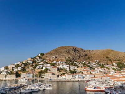 A harbor filled with sailboats and yachts at the base of a steep, arid hill densely covered with white-washed buildings and terracotta roofs under a clear blue sky.