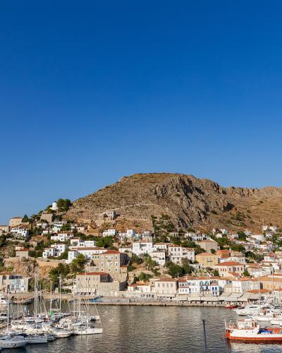 A harbor filled with sailboats and yachts at the base of a steep, arid hill densely covered with white-washed buildings and terracotta roofs under a clear blue sky.