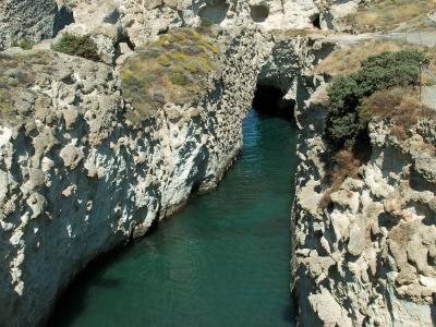 A view looking down into a narrow, deep channel of turquoise water cut through white limestone cliffs, featuring a dark cave opening at the far end and a paved path along the upper right edge.