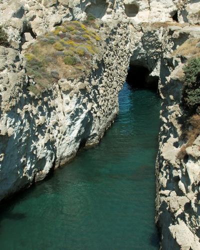 A view looking down into a narrow, deep channel of turquoise water cut through white limestone cliffs, featuring a dark cave opening at the far end and a paved path along the upper right edge.