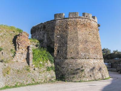 A large, circular stone defensive tower with crenellations and a sloping base, part of a larger fortification wall, set against a clear blue sky with a dirt road and a truck visible in the foreground.