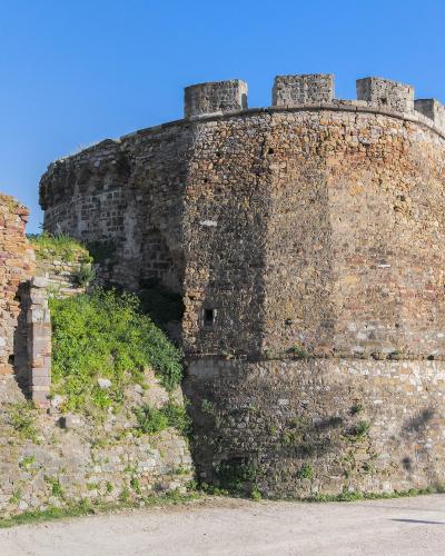 A large, circular stone defensive tower with crenellations and a sloping base, part of a larger fortification wall, set against a clear blue sky with a dirt road and a truck visible in the foreground.