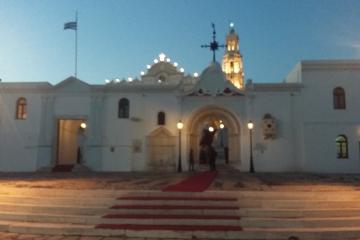 A white-washed church complex with a central arched entrance, a bell tower, and a Greek flag flying on a pole to the left, illuminated at dusk.