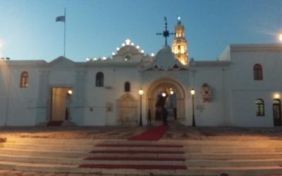 A white-washed church complex with a central arched entrance, a bell tower, and a Greek flag flying on a pole to the left, illuminated at dusk.