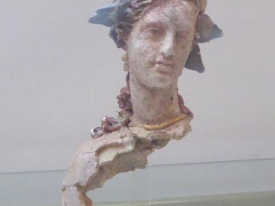 A terracotta figurine of a female head and neck with a headdress, displayed on a clear stand against a plain background.
