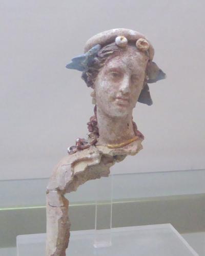 A terracotta figurine of a female head and neck with a headdress, displayed on a clear stand against a plain background.