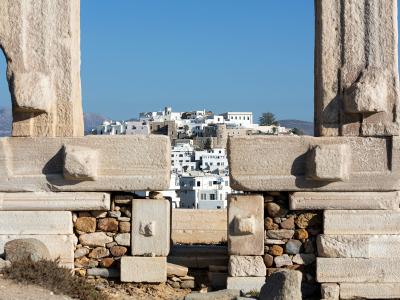 The image shows the massive marble pillars and entablature of the Temple of Apollo at the Portara, with the white-washed buildings of Naxos Chora visible in the background.