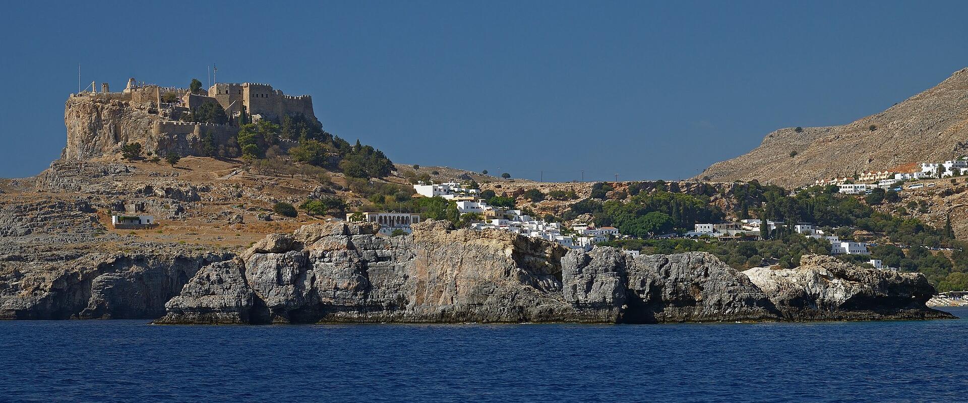 The image shows ancient stone fortifications, crenellated walls, and scattered architectural ruins on a rocky hilltop under a blue sky.