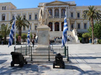 A large neoclassical building with a central statue on a pedestal, flanked by Greek flags and cannons, situated in a paved public square with palm trees.