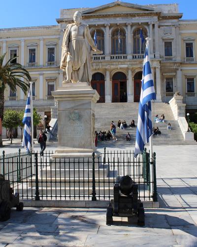 A large neoclassical building with a central statue on a pedestal, flanked by Greek flags and cannons, situated in a paved public square with palm trees.