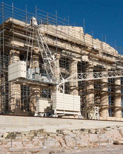 A large ancient Greek temple with Doric columns, heavily covered in scaffolding and construction equipment, indicating active restoration work.