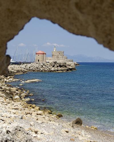 A view through a stone archway of the entrance to the Old Port of Rhodes, featuring the Fort of Saint Peter and the lighthouse on a rocky breakwater.