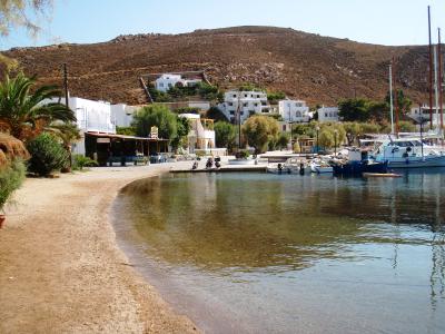 A coastal scene showing a small harbor with white buildings, boats, and a pebbly beach under a hillside.