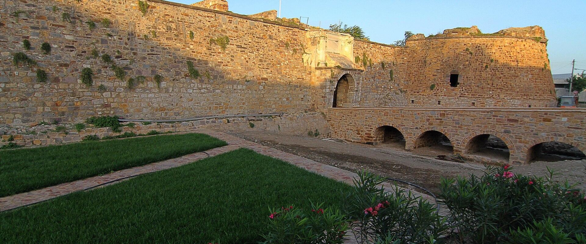 A stone fortress wall with an arched gateway and a Greek flag flying on top, overlooking a grassy courtyard area.