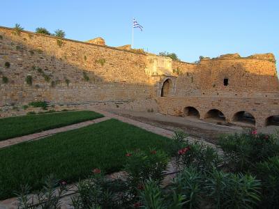 A stone fortress wall with an arched gateway and a Greek flag flying on top, overlooking a grassy courtyard area.