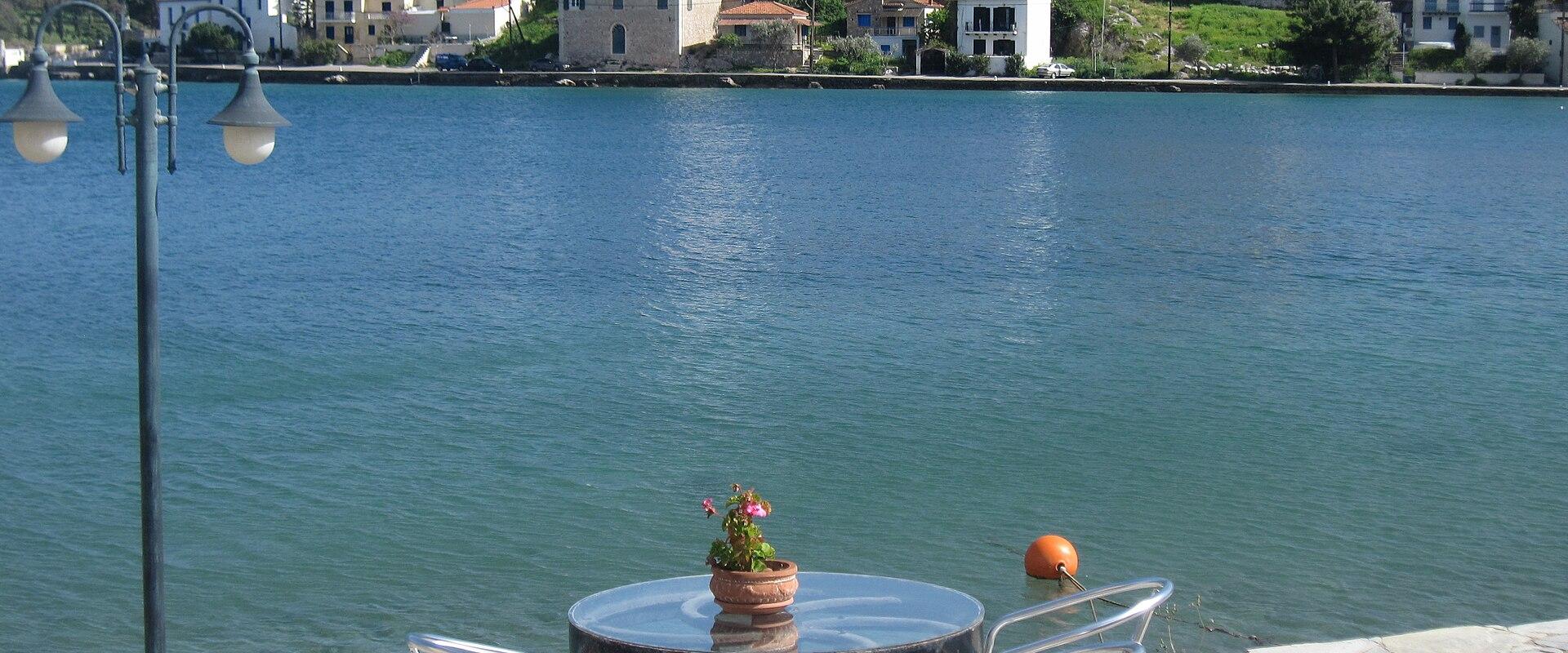 A waterfront view of a Greek coastal town featuring a stone promenade with a table and chairs in the foreground, overlooking a harbor with a hillside village and a prominent church with a dome and bell tower in the background.