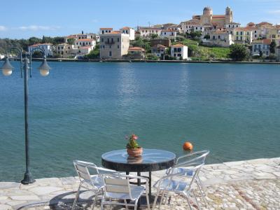 A waterfront view of a Greek coastal town featuring a stone promenade with a table and chairs in the foreground, overlooking a harbor with a hillside village and a prominent church with a dome and bell tower in the background.
