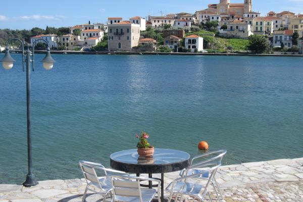 A waterfront view of a Greek coastal town featuring a stone promenade with a table and chairs in the foreground, overlooking a harbor with a hillside village and a prominent church with a dome and bell tower in the background.
