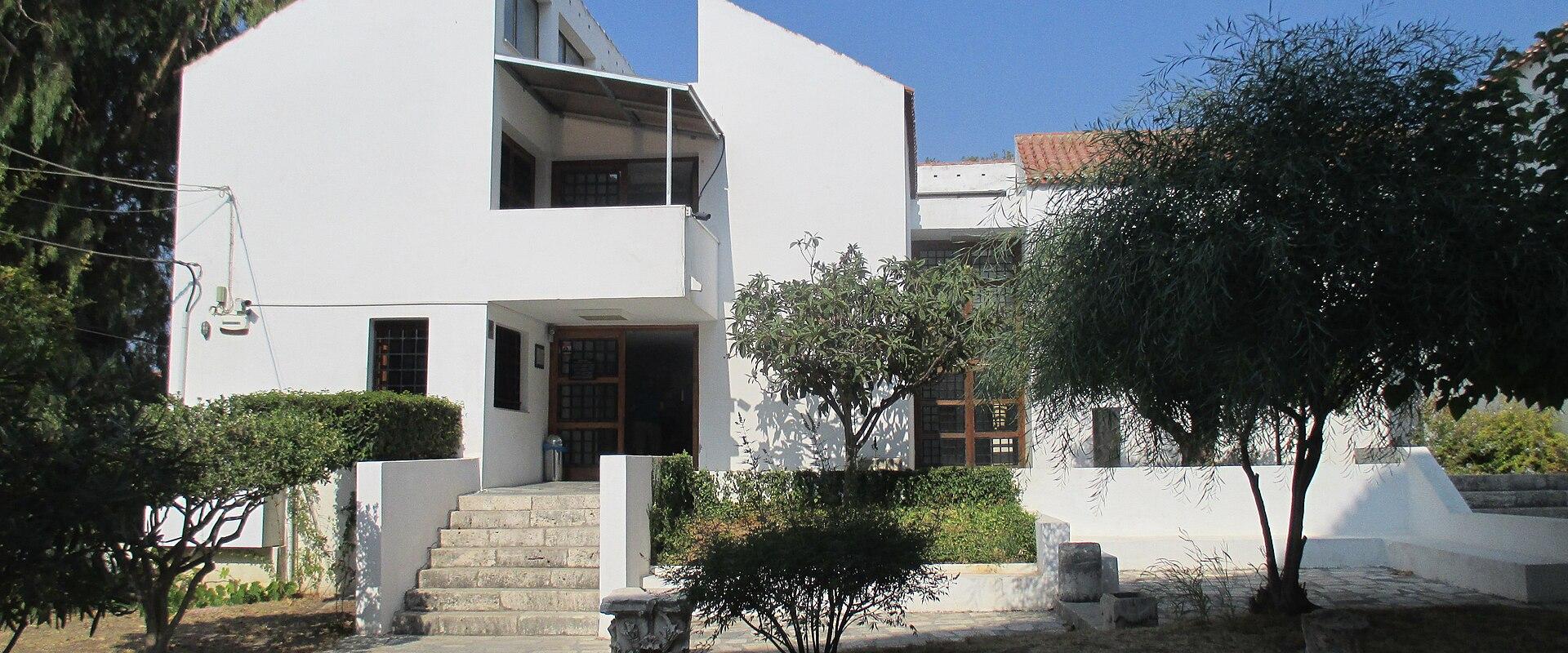 A white, modernist-style building with angular architecture, stone steps leading to an entrance, and a small stone sign in the foreground reading "ΕΙΣΟΔΟΣ / ENTRANCE".