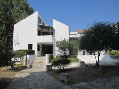 A white, modernist-style building with angular architecture, stone steps leading to an entrance, and a small stone sign in the foreground reading "ΕΙΣΟΔΟΣ / ENTRANCE".