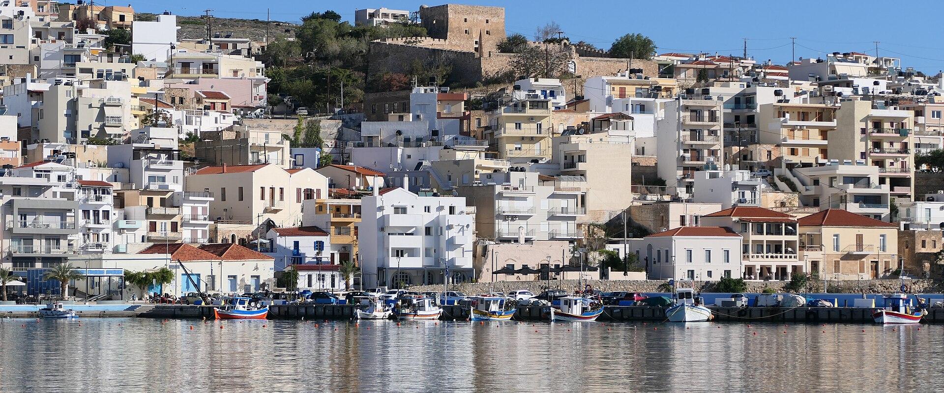 A coastal town with a harbor filled with small fishing boats, backed by a hillside of white and beige buildings, and a stone fortress structure visible at the top of the hill.