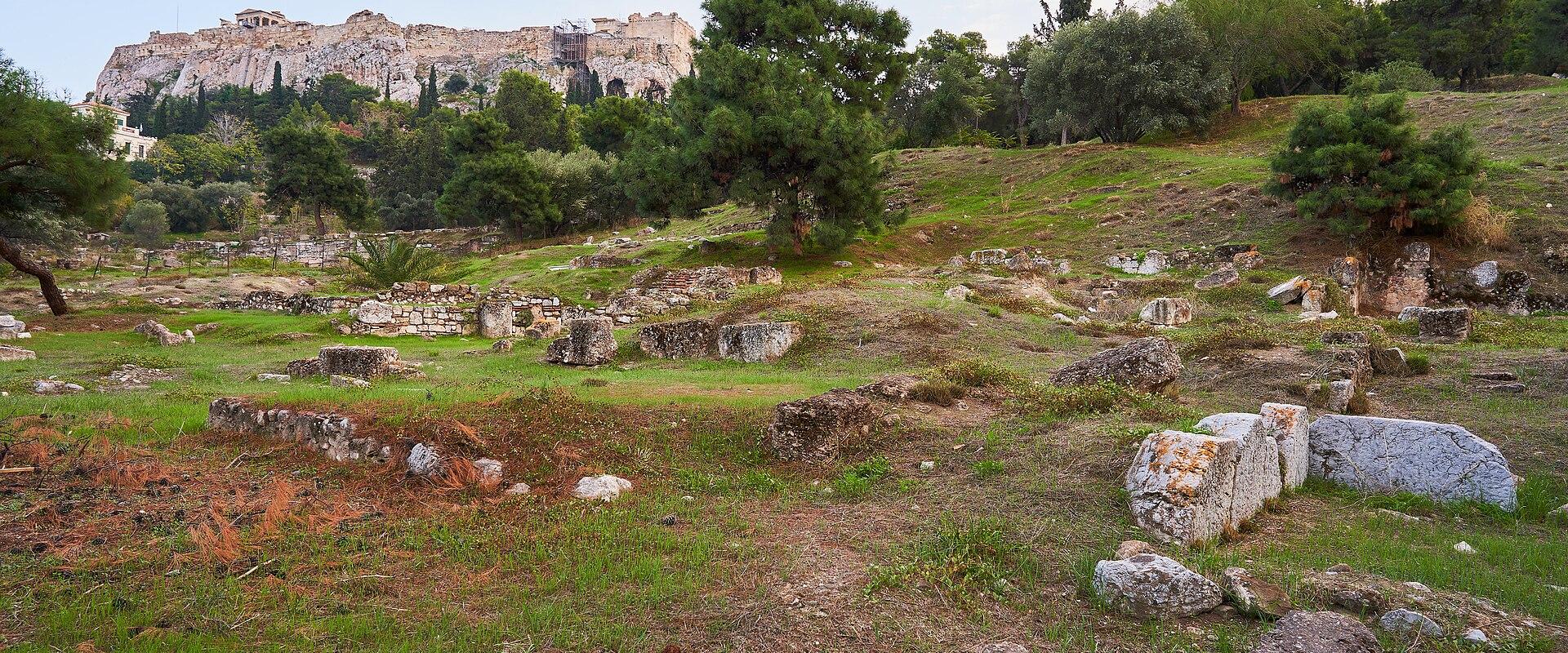 A view of the Acropolis hill and its ruins (including the Parthenon) rising above a grassy foreground scattered with ancient stone foundations and ruins.