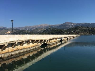 A long, light-colored stone bridge with multiple arches spanning a calm inlet, featuring black lampposts along the walkway and mountains in the background.