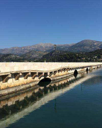 A long, light-colored stone bridge with multiple arches spanning a calm inlet, featuring black lampposts along the walkway and mountains in the background.