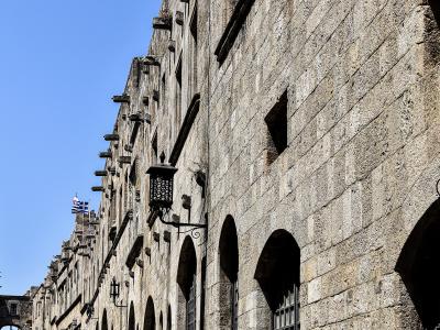 A low-angle view of a long, uniform medieval stone building with a ground-floor arcade of arches and a row of wooden corbels along the roofline, with a Greek flag flying in the distance.
