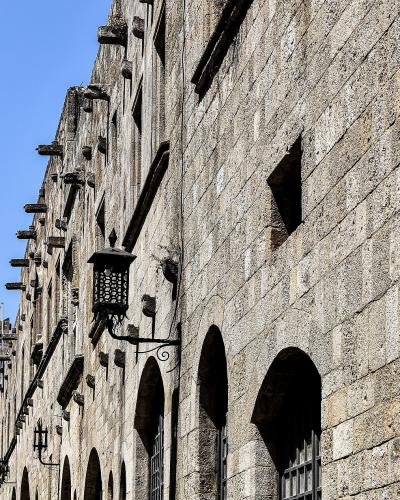 A low-angle view of a long, uniform medieval stone building with a ground-floor arcade of arches and a row of wooden corbels along the roofline, with a Greek flag flying in the distance.